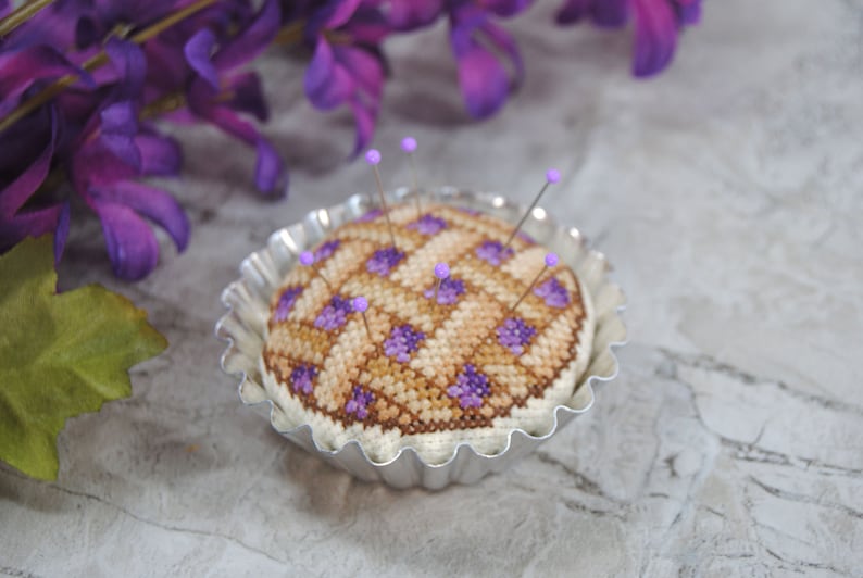 May include: A purple and brown cross-stitch pincushion in the shape of a pie, with purple pins, sitting in a silver pie pan.