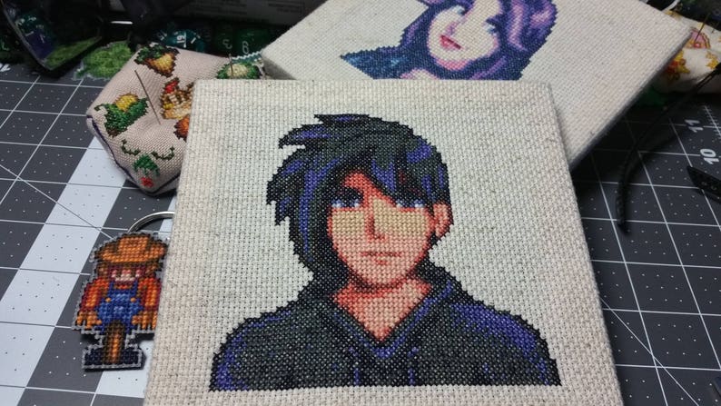 May include: Cross-stitch embroidery of a person with black hair and a blue shirt. The person is looking directly at the viewer.