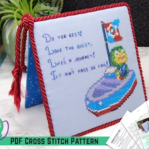 May include: Cross-stitch pattern featuring a boat with a character and a flag, with the text "Do yer best, Love the quest, Life's a journey! It ain't pass or fail!" The design is framed with red cording.