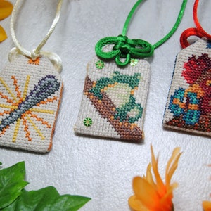 May include: Three embroidered Japanese omamori charms with different designs. One features a microphone with sunburst rays, another a frog on a branch, and the third a bird-like creature. Each charm has a different colored cord.