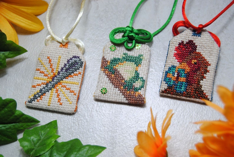 May include: Three embroidered Japanese omamori charms with different designs. One features a microphone with sunburst rays, another a frog on a branch, and the third a bird-like creature. Each charm has a different colored cord.