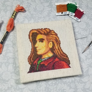 May include: Cross-stitch embroidery of a pixelated character with brown hair and a red jacket. The embroidery is on a white canvas and is surrounded by embroidery floss and scissors.