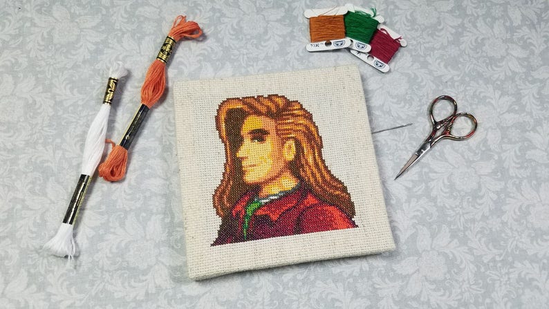 May include: Cross-stitch embroidery of a pixelated character with brown hair and a red jacket. The embroidery is on a white canvas and is surrounded by embroidery floss and scissors.