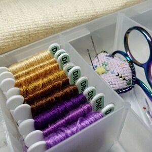May include: A clear plastic storage box with a variety of embroidery floss spools, needles, and a small finished embroidery project. The floss is organized by color, with gold, brown, and purple shades. The embroidery project is a pink heart with a yellow star.