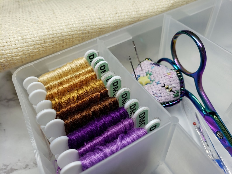May include: A clear plastic storage box with a variety of embroidery floss spools, needles, and a small finished embroidery project. The floss is organized by color, with gold, brown, and purple shades. The embroidery project is a pink heart with a yellow star.