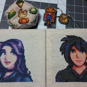 May include: Two cross-stitch embroidery pieces on beige fabric. The left piece features a purple-haired female character with blue eyes. The right piece features a male character with black hair and blue eyes. Both characters are wearing blue clothing.