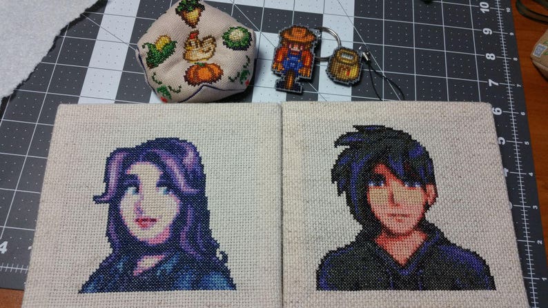 May include: Two cross-stitch embroidery pieces on beige fabric. The left piece features a purple-haired female character with blue eyes. The right piece features a male character with black hair and blue eyes. Both characters are wearing blue clothing.
