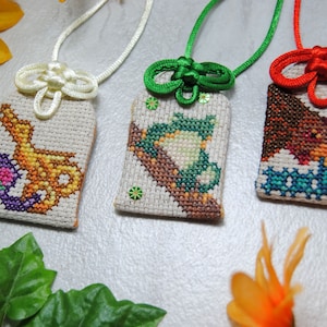 May include: Three handmade cross-stitch pendants with different designs. One features a yellow and purple design, another a green frog, and the last a brown bird. Each pendant is attached to a cord with a decorative knot.