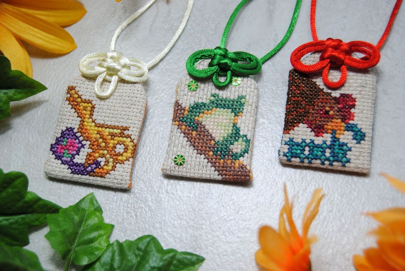 May include: Three handmade cross-stitch pendants with different designs. One features a yellow and purple design, another a green frog, and the last a brown bird. Each pendant is attached to a cord with a decorative knot.