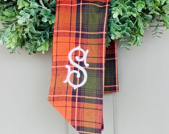 May include: A fall-themed wreath with a plaid ribbon featuring the embroidered letter "S". The ribbon has orange, green, and brown tones. The wreath is adorned with green foliage, creating a welcoming seasonal decoration.