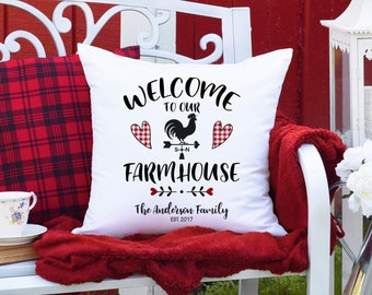 May include: White square pillow with the text "WELCOME TO OUR FARMHOUSE" and a rooster weathervane design. A red and black plaid pillow is next to it. The bench has a red blanket and a teacup.