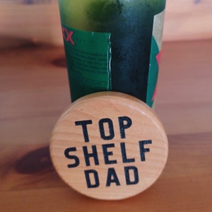 May include: A wooden coaster with the text "Top Shelf Dad" in black lettering.