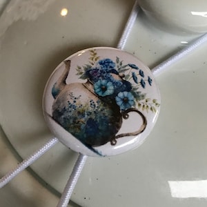 May include: White ceramic teapot with a white string and a blue and white button with a floral design of a teapot with blue flowers.