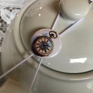 May include: A white ceramic teapot with a white elastic band and a round button featuring a vintage pocket watch with Roman numerals.