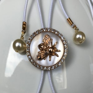 May include: A white and gold necklace with a bee charm surrounded by rhinestones. The necklace has two white pearl accents.