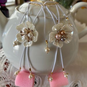 May include: Two white flower-shaped pendants with pearl and rhinestone accents hang from white cords. The pendants are on a white teapot.