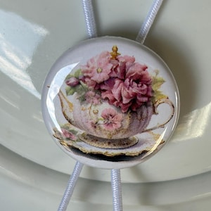 May include: A round, white button with a floral design featuring a teapot and pink flowers. The button is attached to a white cord. The design is a vintage style, with a focus on the floral arrangement.