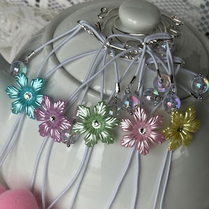 May include: Six iridescent flower tea bag charms with white elastic cords and silver clasps. The charms are hanging from a silver teapot lid holder.