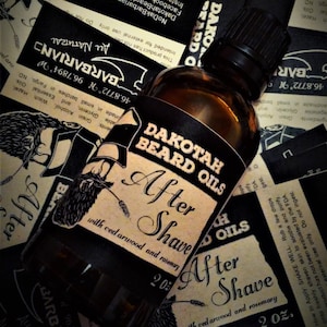 May include: A brown glass bottle with a black label that reads "Dakotah Beard Oils After Shave with cedarwood and rosemary 2 oz.". The bottle is sitting on a black and white background.