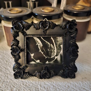 Puede incluir: Un pequeño marco de fotos negro adornado con una imagen en blanco y negro. El marco tiene un diseño floral detallado. Detrás del marco hay tres tarros con tapas negras y detalles dorados. El fondo es una superficie texturizada de color claro.