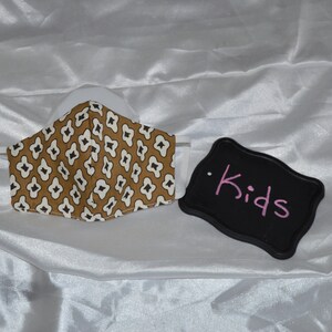 May include: A brown and white patterned face mask with a black elastic band. A black sign with the word "Kids" written in pink is next to the mask.