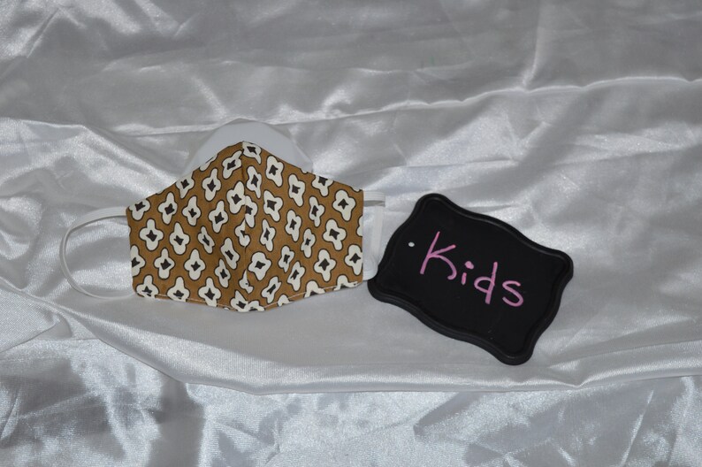 May include: A brown and white patterned face mask with a black elastic band. A black sign with the word "Kids" written in pink is next to the mask.