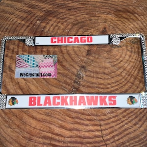 May include: A silver license plate frame with rhinestones around the border. The frame has a white background with the text "CHICAGO BLACKHAWKS" in red and black lettering. The frame also features a small blackhawk logo on each side.