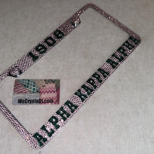 May include: A rectangular license plate frame covered in light pink rhinestones. The top bar features the number 1908 in dark green rhinestones, while the bottom bar displays the text ALPHA KAPPA ALPHA in dark green rhinestones.