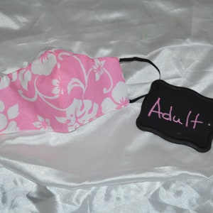 Pu&ograve; includere: Maschera facciale rosa con un motivo floreale bianco. La maschera ha lacci elastici neri per le orecchie. Un cartello nero in ardesia con la parola "Adulto" scritta in rosa.