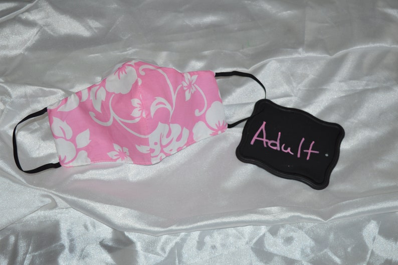 Pu&ograve; includere: Maschera facciale rosa con un motivo floreale bianco. La maschera ha lacci elastici neri per le orecchie. Un cartello nero in ardesia con la parola "Adulto" scritta in rosa.