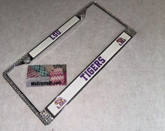 May include: A rectangular license plate frame featuring a white background with purple text reading LSU and TIGERS. The frame is bordered by clear, reflective rhinestones and includes two small LSU logos. A small rectangular card reads WeCrystalit.com.