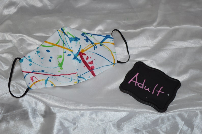 May include: White face mask with a colorful paint splatter design. The mask has black elastic ear loops. A black sign with the word "Adult" written in pink is in the image.