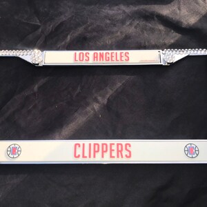 May include: A silver license plate frame with rhinestones around the border. The frame has two white license plates with red text. The top plate says "LOS ANGELES" and the bottom plate says "CLIPPERS".