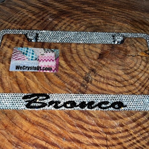 May include: A silver license plate frame with a black "Bronco" inscription and a clear crystal design. The frame is on a wooden surface.