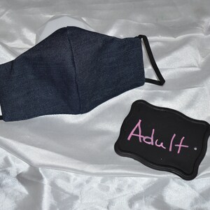 Pu&ograve; includere: Una maschera facciale in denim blu scuro con elastici per le orecchie neri. Uno sfondo nero con la parola "Adulto" scritta con gesso rosa.