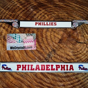 May include: A silver license plate frame with rhinestones. The frame has a white insert with the words "PHILLIES" and "PHILADELPHIA" in red lettering. The frame also has a small Phillies logo on each side.