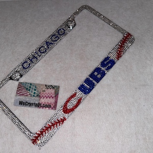 May include: A rectangular license plate frame covered in clear rhinestones. The top features the word CHICAGO in blue rhinestones, while the right side displays the word CUBS in blue rhinestones, accented by a red rhinestone baseball stitch design.