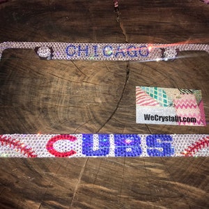 May include: A white, blue, and red rhinestone license plate frame with the words "CHICAGO CUBS" and a baseball design. The frame is on a wooden surface.