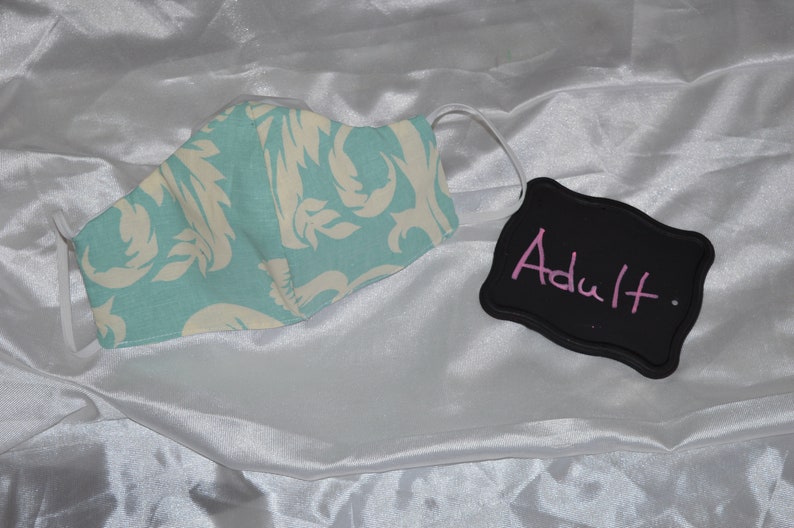 Pu&ograve; includere: Una maschera facciale blu chiaro con un motivo floreale bianco. La maschera &egrave; etichettata "Adulto" su una lavagna nera.