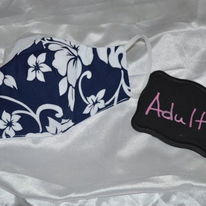 Pu&ograve; includere: Una maschera facciale blu e bianca a fiori con sfondo bianco e un motivo floreale blu navy. La maschera &egrave; etichettata "Adulto" su una lavagna nera.