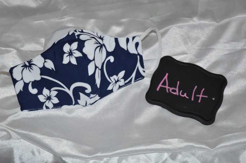 Pu&ograve; includere: Una maschera facciale blu e bianca a fiori con sfondo bianco e un motivo floreale blu navy. La maschera &egrave; etichettata "Adulto" su una lavagna nera.