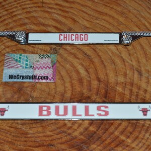 May include: A silver license plate frame with rhinestones around the border. The frame has a white background with the words "CHICAGO BULLS" in red lettering. A red bull logo is on each side of the text.