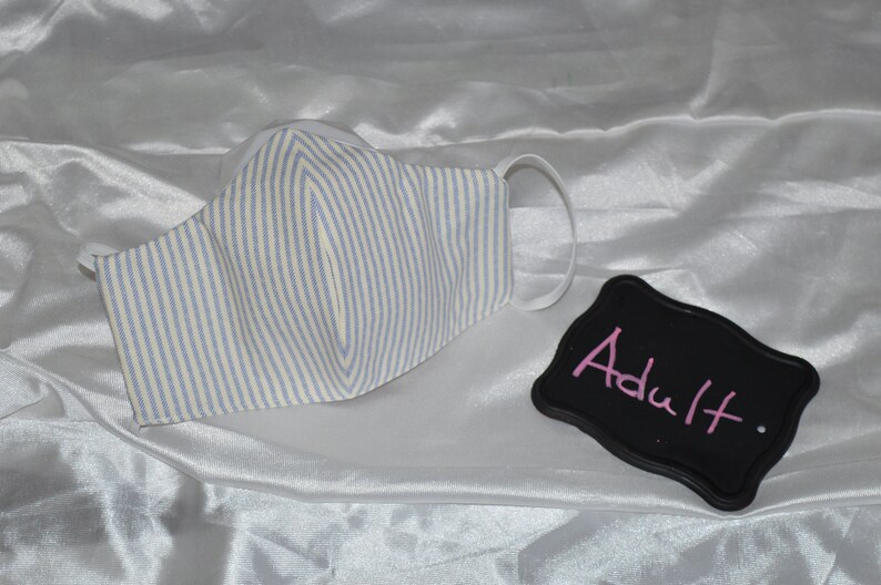 May include: White face mask with blue and white stripes. The mask has elastic ear loops. A black sign with the word "Adult" written in pink is in the image.
