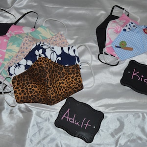 Pu&ograve; includere: Assortimento di maschere per il viso in tessuto con vari motivi e colori, tra cui stampa leopardata, floreale e blu con pois bianchi. Le maschere sono disposte in due gruppi, etichettati "Adult" e "Kids".