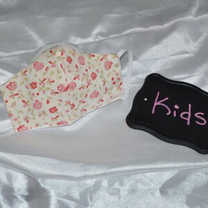May include: A white face mask with a pink floral pattern. The mask has elastic ear loops. A black sign with the word "Kids" written in pink is in the background.