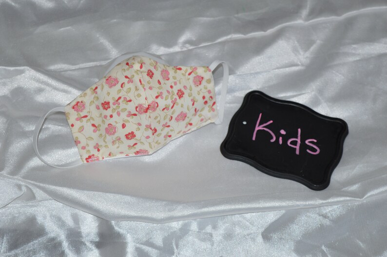 May include: A white face mask with a pink floral pattern. The mask has elastic ear loops. A black sign with the word "Kids" written in pink is in the background.