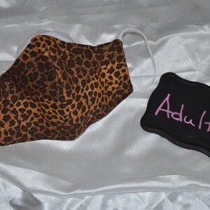 Pu&ograve; includere: Una maschera facciale in tessuto con stampa leopardata marrone e nera con elastici per le orecchie bianchi. Un cartello nero in ardesia con la parola "Adulto" scritta in rosa.