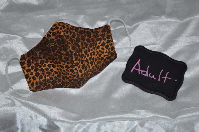 Pu&ograve; includere: Una maschera facciale in tessuto con stampa leopardata marrone e nera con elastici per le orecchie bianchi. Un cartello nero in ardesia con la parola "Adulto" scritta in rosa.