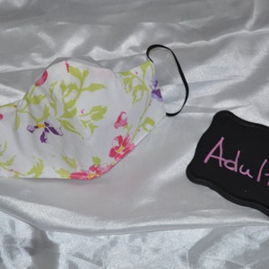 May include: White face mask with a floral pattern. The mask has black elastic ear loops. A black sign with the word "Adult" written in pink is in the background.