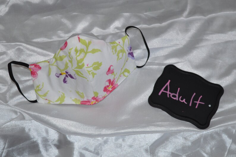 May include: White face mask with a floral pattern. The mask has black elastic ear loops. A black sign with the word "Adult" written in pink is in the background.